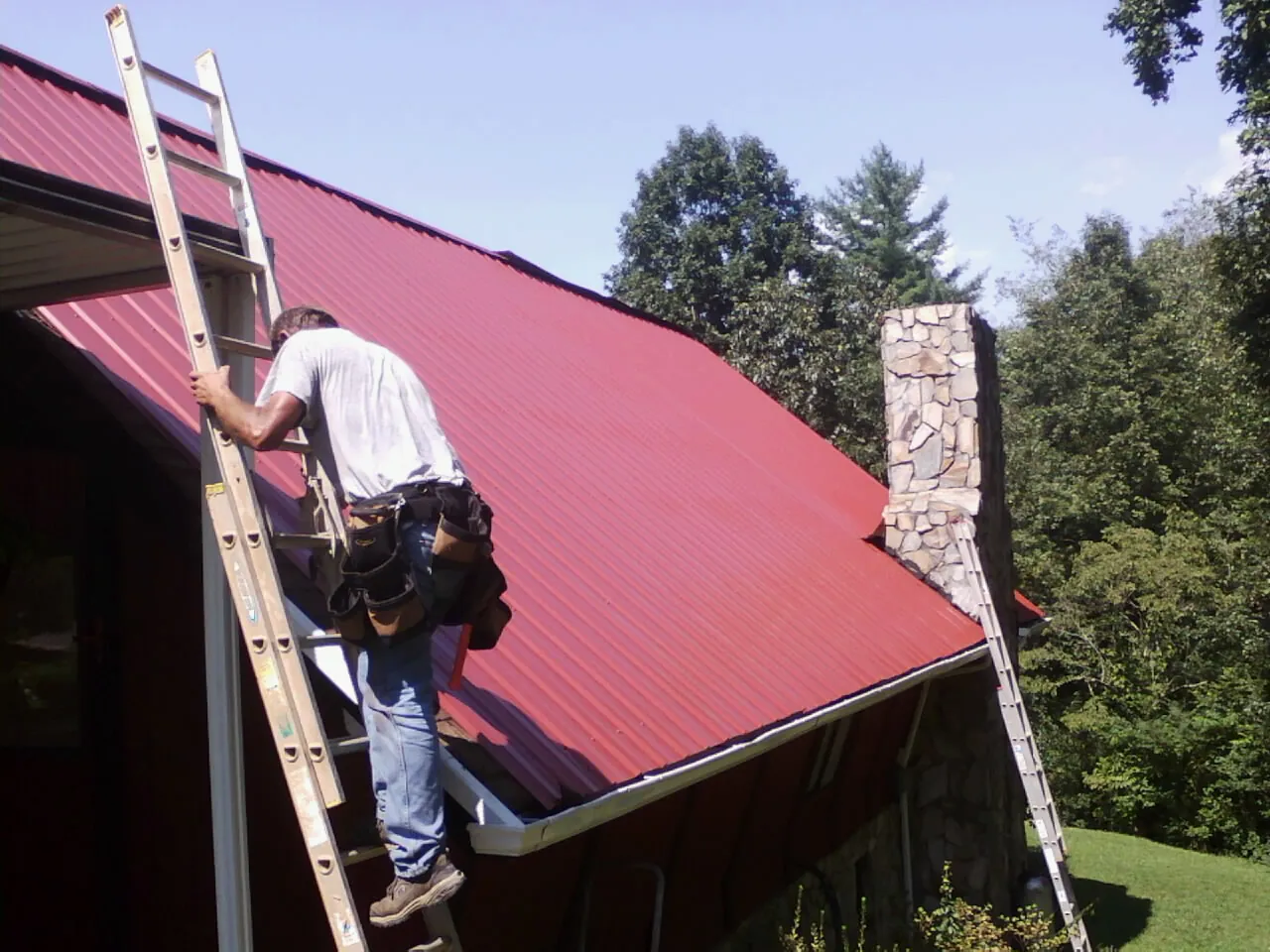 Roofer on a ladder beside a red metal roof and stone chimney — Soffit & Fascia Repair by Roofing Brothers in Colfax